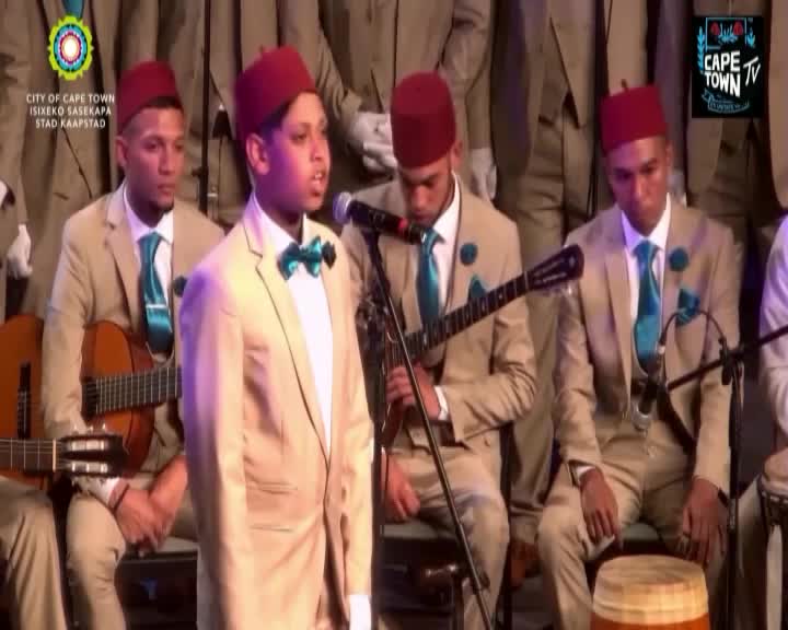 A young man in a tan suit and red fez sings into a microphone, his teal bowtie catching the light. To his left, another man in similar attire strums a guitar, while others in the ensemble, also in tan suits and red fezzes, are visible behind them. The City of Cape Town logo is displayed on a screen behind the performers.