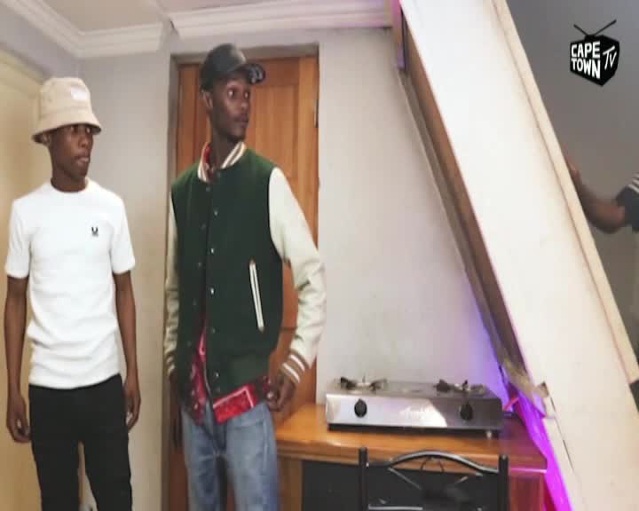 Two young men stand in a room, one in a white t-shirt and bucket hat, the other in a green varsity jacket. A large white panel is being tilted into place behind them, obscuring a gas stove.
