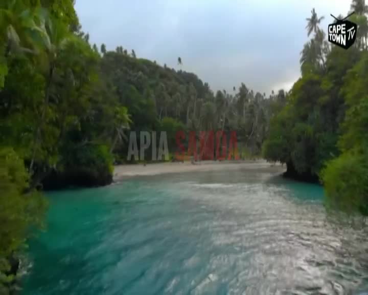 The turquoise water laps the shore of a tropical beach, where large red letters spell out "SAMOA." Lush green foliage crowds the coastline under a cloudy sky.