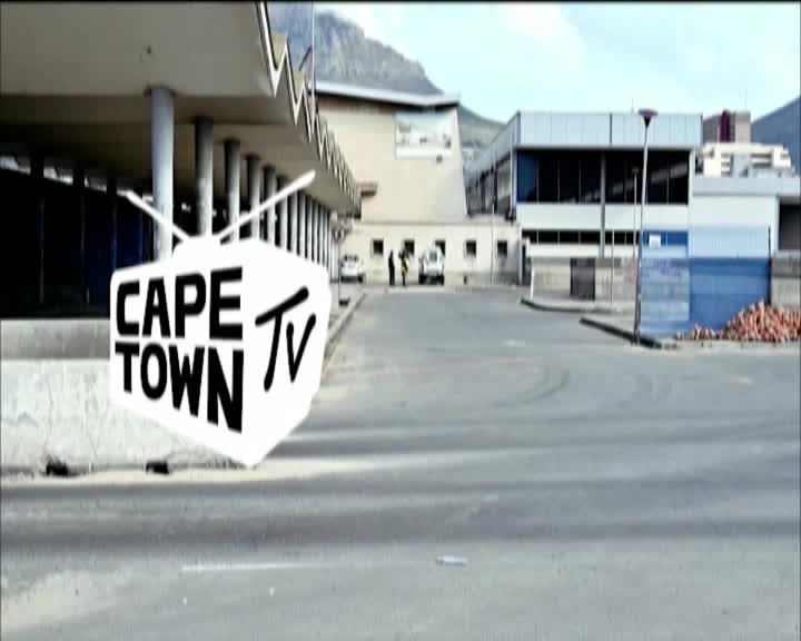 A white van is parked on the street, and two people stand beside it, looking towards the buildings. A large, white television graphic with "CAPE TOWN TV" printed on it floats in the foreground.