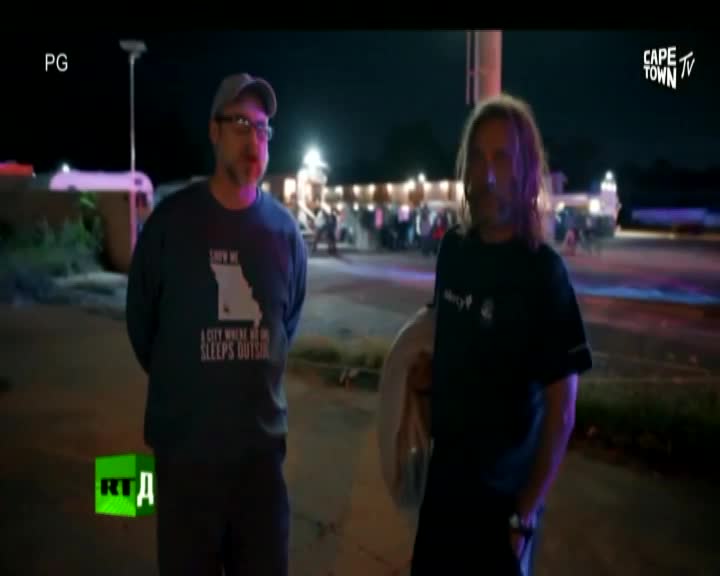 Two men stand in the dark, illuminated by flashing lights in the background. A television crew's equipment is visible, suggesting a news report is underway.