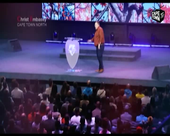 A man in a rust-colored shirt stands at a podium on a stage, addressing a seated congregation. The backdrop displays a vibrant, abstract image, and the Cape Town TV logo is visible in the corner. A man in a rust-colored shirt stands at a podium on a stage, addressing a seated congregation. The backdrop displays a vibrant, abstract image, and the Cape Town TV logo is visible in the corner.