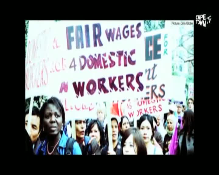 A large banner demanding "FAIR WAGES FOR DOMESTIC WORKERS" is held aloft by a crowd of people. The faces in the foreground are a mix of determination and hope as they stand together. A large banner demanding "FAIR WAGES FOR DOMESTIC WORKERS" is held aloft by a crowd of people. The faces in the foreground are a mix of determination and hope as they stand together.