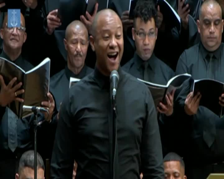 The lead singer's mouth is wide open, belting out a note as the rest of the choir, dressed in black, holds their songbooks open. This performance, broadcast on Cape Town TV, showcases the powerful voices of this South African ensemble. The lead singer's mouth is wide open, belting out a note as the rest of the choir, dressed in black, holds their songbooks open. This performance, broadcast on Cape Town TV, showcases the powerful voices of this South African ensemble.