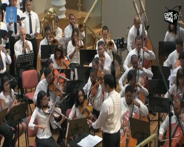 The conductor gestures with his left hand as the orchestra plays. Violins and cellos fill the stage, their bows moving in unison. The conductor gestures with his left hand as the orchestra plays. Violins and cellos fill the stage, their bows moving in unison.
