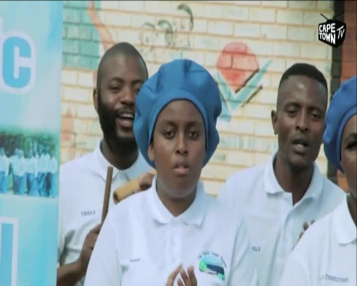 A group of people in white shirts and blue headwear sing with passion. A banner with the Cape Town TV logo is visible in the background. A group of people in white shirts and blue headwear sing with passion. A banner with the Cape Town TV logo is visible in the background.