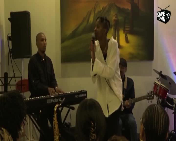 A woman in a white shirt sings into a microphone, her hands gesturing. Beside her, a man plays a Roland keyboard, and a guitarist strums his instrument. A woman in a white shirt sings into a microphone, her hands gesturing. Beside her, a man plays a Roland keyboard, and a guitarist strums his instrument.