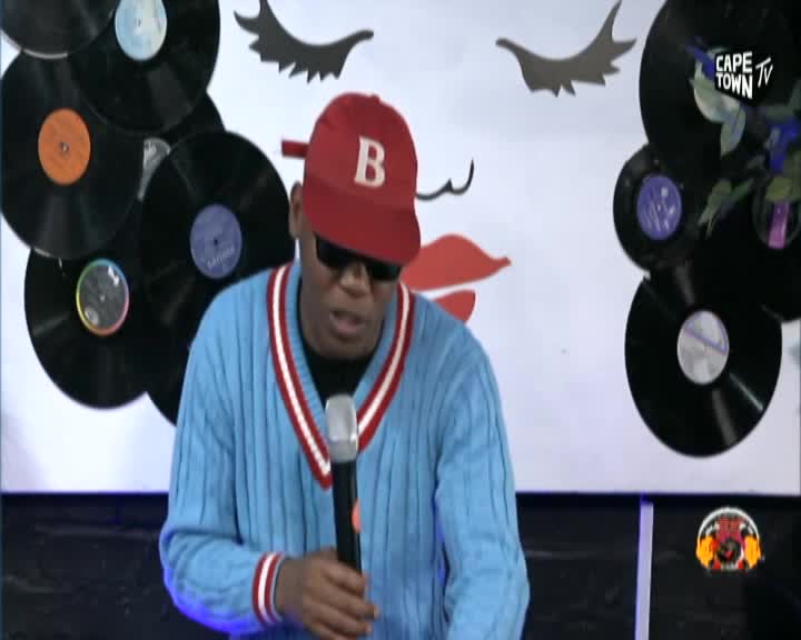 A man in a red cap and blue sweater holds a microphone, speaking into it. He's on a set decorated with vinyl records and a large graphic of lips, likely for a Cape Town TV broadcast.