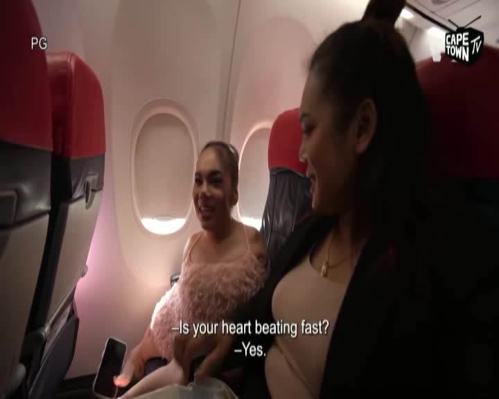 Two women sit in airplane seats, illuminated by the soft glow of the cabin lights. One woman, wearing a fluffy pink top, smiles as she holds a phone. The other woman, in a dark jacket, leans in, her expression friendly.