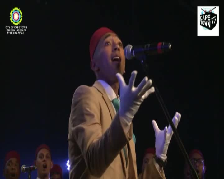 A man in a tan suit and red fez sings into a microphone, his white-gloved hands gesturing expressively. Behind him, a chorus of performers in similar red hats watches intently.