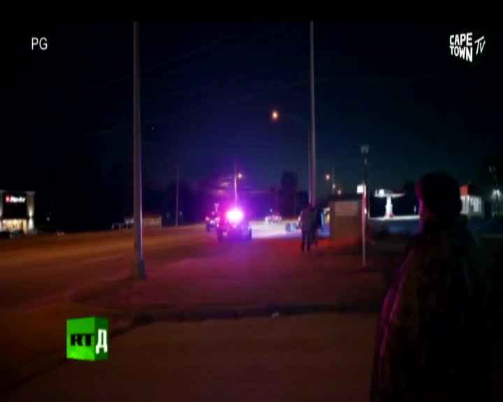 Flashing blue and red lights paint the night sky as a police car idles near a roadside establishment. A few figures stand near the vehicle, their faces obscured by the darkness and the glare of the lights.