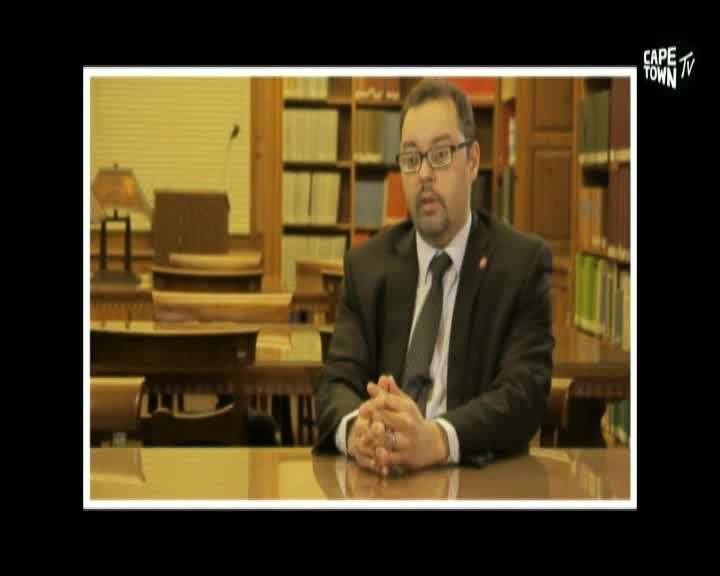 A man in a suit sits with his hands clasped, speaking in what appears to be a library setting. The Cape Town TV logo is visible in the upper right corner.