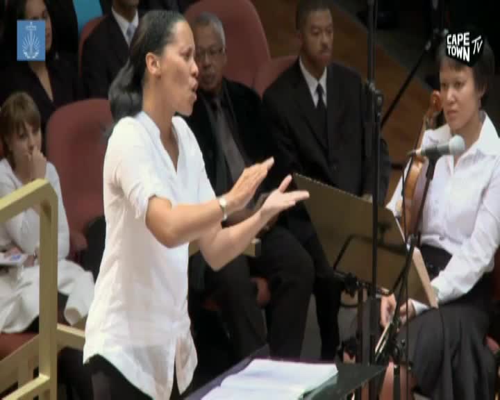 The conductor gestures with open hands, her white shirt crisp against the dark suits of the orchestra behind her. A violinist to her right, also in white, holds her bow steady.
