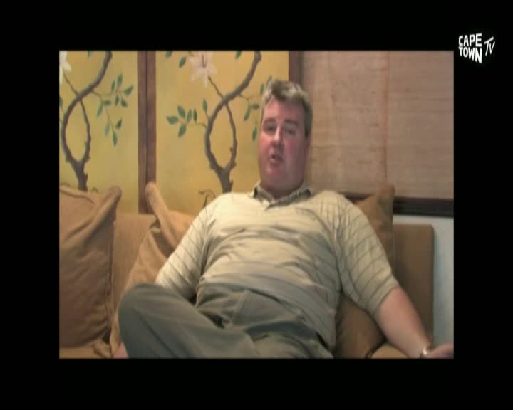 A man in a striped polo shirt sits on a couch, his legs crossed. The background features a warm, patterned wall, and a Cape Town TV logo appears in the corner.
