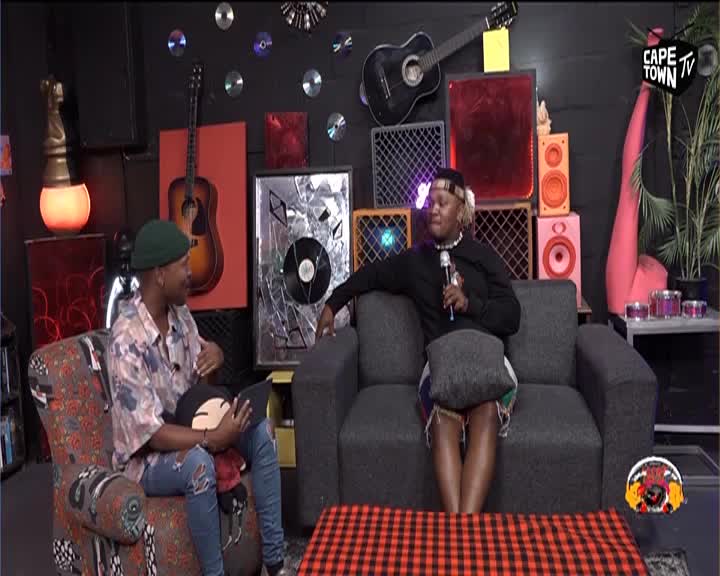 Two people are seated in a studio, one on a patterned armchair and the other on a dark grey couch. The person on the couch holds a microphone, speaking into it as they look towards the person opposite them. The backdrop is decorated with guitars, vinyl records, and colourful speakers, hinting at a music-focused broadcast from Cape Town TV.