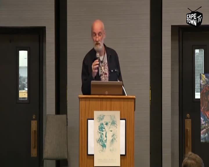 A man with a beard stands at a podium, speaking into a microphone. A laptop rests on the podium in front of him, and a poster with abstract art is displayed on its side. The Cape Town TV logo is visible in the upper right corner.