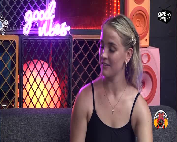 A young woman with blonde hair is speaking, her gaze directed slightly off-camera. Behind her, a neon sign spells out "good vibes" in bright pink, and a large speaker with a pink cone sits stacked on another orange one.