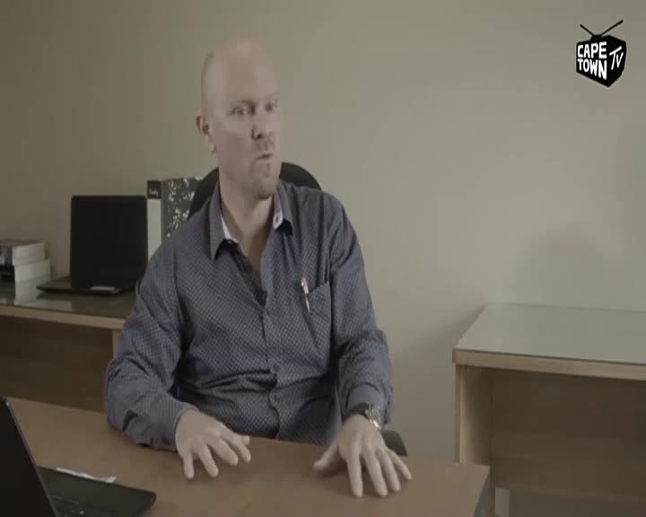 A man in a patterned blue shirt gestures with his hands as he speaks at a desk. A Cape Town TV logo appears in the corner of the frame.