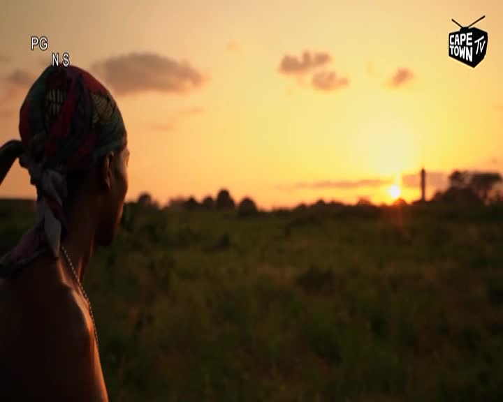 The sun dips low over the South African landscape, casting a warm glow across the fields. A man, his head wrapped in a patterned cloth, turns to watch the Cape Town TV broadcast as the day ends.