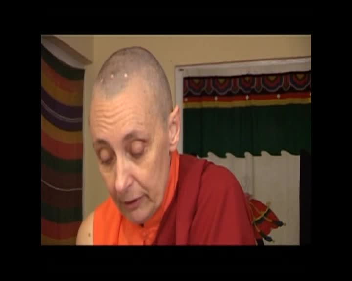 A person with a shaved head and wearing orange and red robes looks down, their eyes closed. Behind them, a colorful tapestry hangs on the wall.