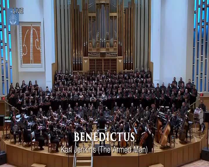A massive choir fills the stage behind an orchestra, all bathed in the warm glow of stage lights. The conductor, a solitary figure in black, directs the ensemble as they perform Karl Jenkins' "Benedictus."