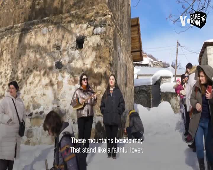 A woman plays a stringed instrument, her voice rising over the quiet hum of a Cape Town TV crew. Snow blankets the ground around a group of onlookers gathered near a stone building.