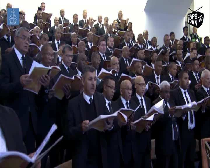 A large choir, dressed in black suits and ties, stands in rows, holding open songbooks. The men's faces are turned upwards, their mouths open as they sing. A Cape Town TV logo is visible in the upper right corner.