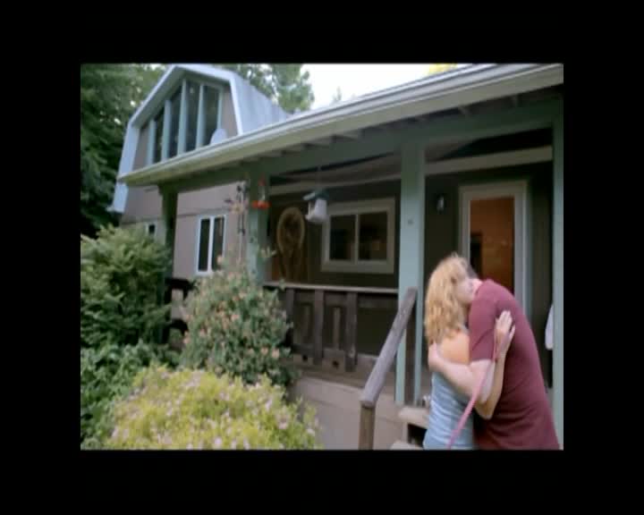 A man and woman embrace on a porch, their heads tilted together. The late afternoon sun casts long shadows across the lawn.