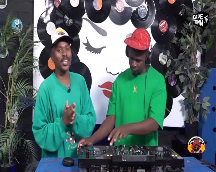 Two men stand before a DJ setup, one talking animatedly while the other focuses on the turntables. The backdrop features a large graphic of a woman's face surrounded by vinyl records, a familiar sight on Cape Town TV.