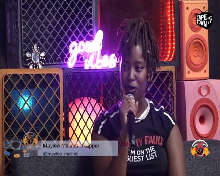 A young woman with dreadlocks holds a microphone, speaking into it. Behind her, a neon sign glows "good vibes" above a collection of speakers.