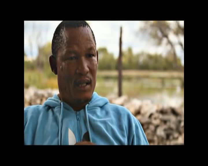 A man in a light blue hoodie speaks, his gaze directed slightly off-camera. Behind him, a blurred landscape suggests an outdoor setting near water.
