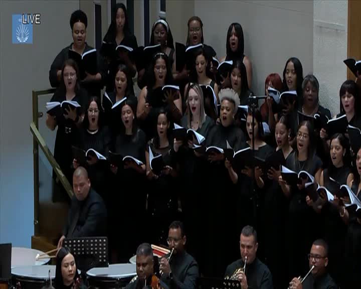 A large choir, dressed in black, sings from open books. Below them, musicians play brass instruments and percussion.