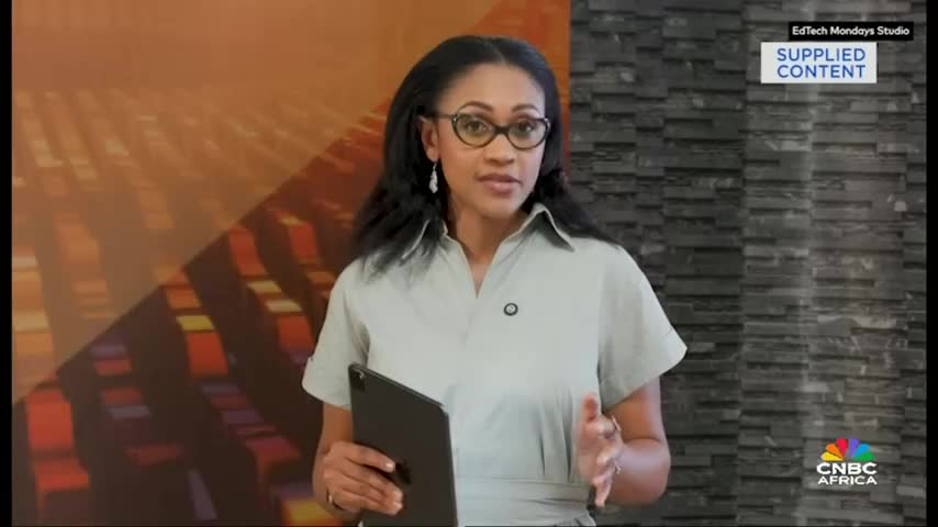 A woman on CNBC Africa, holding a tablet, gestures with her hands as she speaks. The backdrop features a brick wall and a colorful, abstract design.
