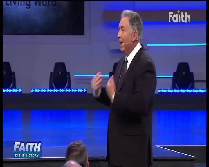 A man in a dark suit gestures as he speaks, his hands moving to emphasize his words on the Faith TV stage. The backdrop is lit with blue lights, and the Faith logo is visible in the corner.
