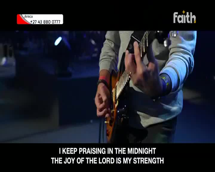 A guitarist on Faith TV, broadcasting from South Africa, strums the strings of his instrument. His fingers dance over the fretboard as the words "I keep praising in the midnight" appear on the screen.
