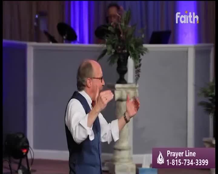 The man in the vest gestures with both hands, his fingers splayed as he speaks on Faith TV. A flower arrangement sits behind him, and the Prayer Line number is visible at the bottom.
