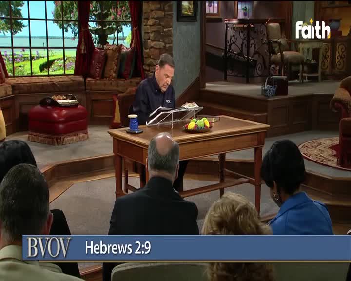 A man in a dark shirt sits at a wooden desk, reading from a book on a stand, with a small audience in front of him. The scene is set in a studio, likely for Faith TV, with a large window and decorative furnishings.
