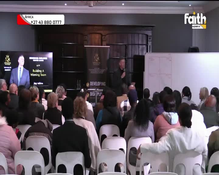 A man on stage, speaking into a microphone, addresses a seated audience. The Faith TV logo is visible, and a banner advertises a business seminar in South Africa.
