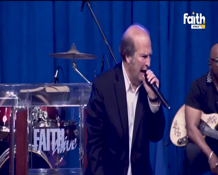 The man in the suit is singing into a microphone, his mouth open wide. A drummer and guitarist are set up behind him, and the Faith TV logo is visible in the corner.
