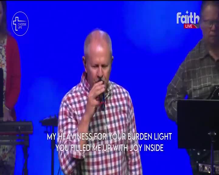 A man in a checkered shirt sings into a microphone on a stage with a blue backdrop, as seen on Faith TV. The lyrics "My Heaviness for your burden light, you filled me up with joy inside" appear on the screen.

