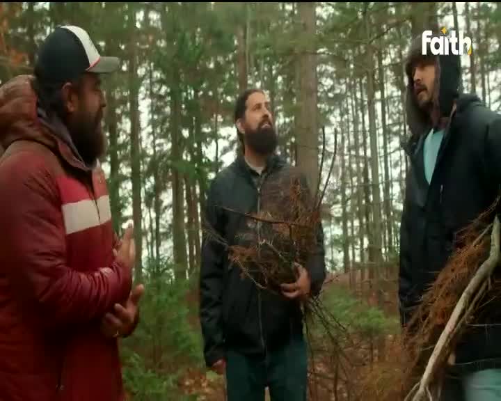 Three men stand in a forest, the one on the left gesturing with his hands. The middle man holds a bundle of branches, while the man on the right looks on, bundled in a coat.
