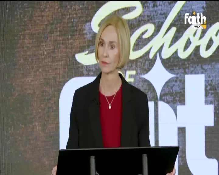 A woman in a black blazer and red shirt stands behind a lectern, speaking. The backdrop behind her features the "Faith Africa" logo and the words "School of..." in a large, stylized font.
