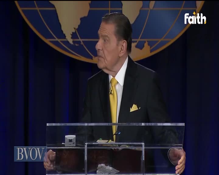 A man in a dark suit and yellow tie gestures slightly as he speaks from behind a clear podium. The Faith TV logo sits in the corner, above a globe graphic.
