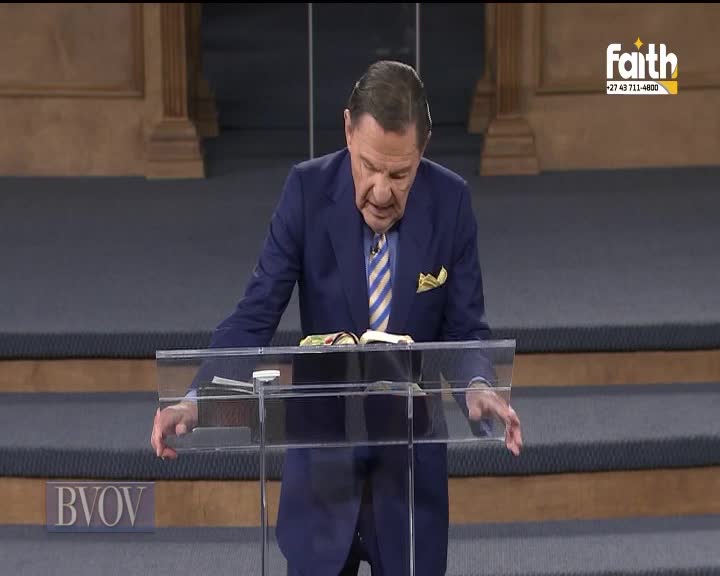 A man in a blue suit leans over a clear podium, his hands resting on it. He's reading from an open book, likely on Faith TV in South Africa.
