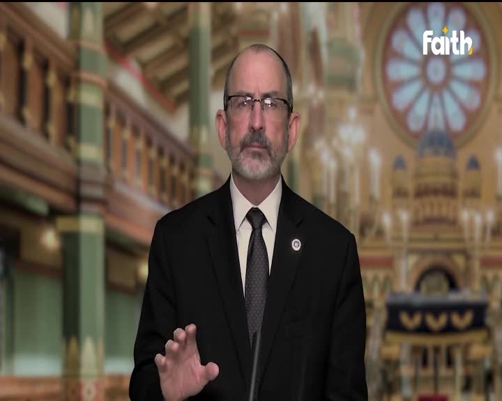 A man in a dark suit gestures with his left hand while speaking. Behind him, a blurred backdrop suggests a church or synagogue, with the "faith" logo visible in the corner.
