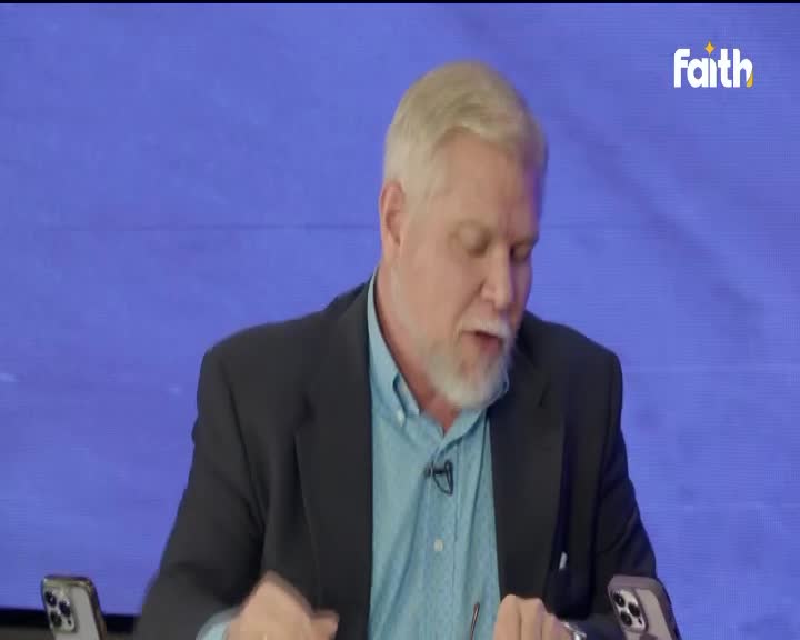 A man with a white beard is speaking, his mouth open as he looks down.  Two phones sit on the table in front of him, and the Faith TV logo is in the corner.
