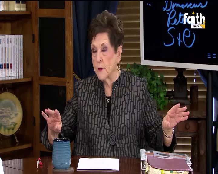 The woman gestures with both hands as she speaks, her patterned jacket prominent against the backdrop. A screen behind her displays "Pet Faith Africa" and some handwritten text.

