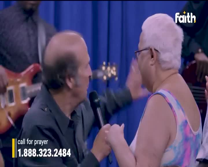 On the Faith TV set, a man in a black shirt holds a microphone, speaking intently to a woman in a tie-dye top. The woman's hand is raised, as if in response, and the band in the background continues to play.
