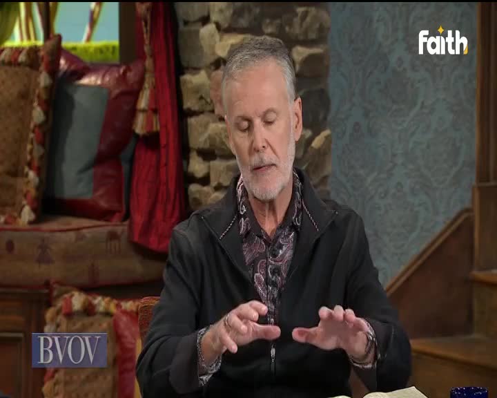 A man with gray hair gestures with his hands, speaking on Faith TV. He's sitting at a table with a book in front of him, likely broadcasting from South Africa.
