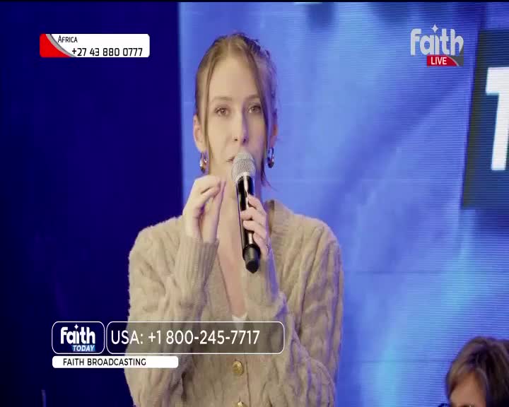 On the Faith Today set, a woman in a beige cardigan holds a microphone, gesturing with her fingers. The screen behind her is a bright blue, and contact information for both South Africa and the USA is displayed.
